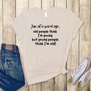 Puede incluir: Una camiseta beige con texto negro que dice "Estoy en una edad extra&ntilde;a... la gente mayor piensa que soy joven. pero la gente joven piensa que soy viejo!"