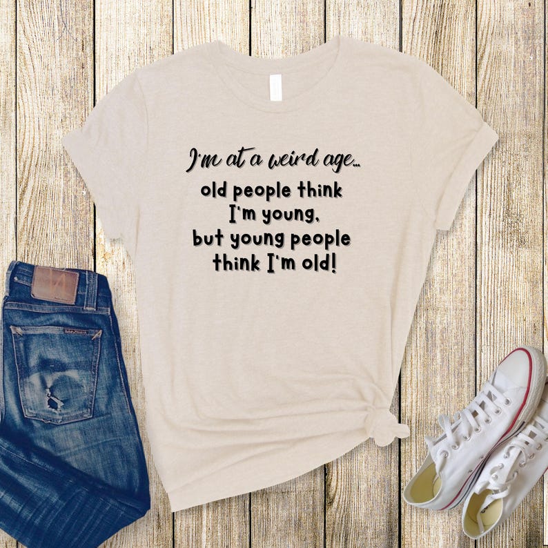 Puede incluir: Una camiseta beige con texto negro que dice "Estoy en una edad extra&ntilde;a... la gente mayor piensa que soy joven. pero la gente joven piensa que soy viejo!"