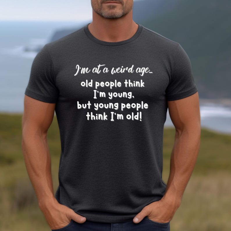Puede incluir: Una camiseta gris oscura con texto blanco que dice "Estoy en una edad extra&ntilde;a... la gente mayor piensa que soy joven, pero la gente joven piensa que soy viejo!"