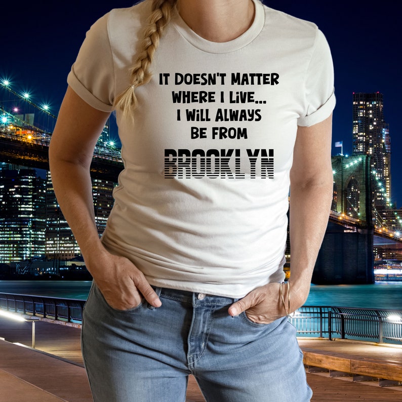 May include: White t-shirt with black text that reads "It doesn't matter where I live... I will always be from BROOKLYN". The shirt is being worn by a person standing in front of a city skyline at night.