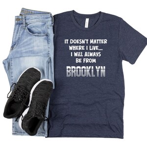 May include: A navy blue t-shirt with white text that reads "It doesn't matter where I live... I will always be from BROOKLYN". The text is in a retro style font with white stripes.