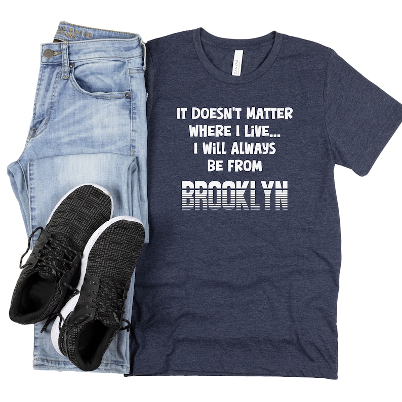 May include: A navy blue t-shirt with white text that reads "It doesn't matter where I live... I will always be from BROOKLYN". The text is in a retro style font with white stripes.