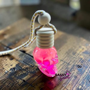 Könnte beinhalten: Ein hängender Auto-Lufterfrischer mit einer durchscheinenden neonpinken, totenkopfförmigen Glasflasche, einem hellen Holzdeckel und einer runden Holzperle. Eine geflochtene Kordel mit gelben und weißen Mustern ist befestigt. Der Text SASSY Saint ist sichtbar.
