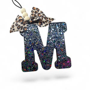 May include: A letter M ornament covered in black and multi-colored glitter. The letter features a leopard print fabric bow at the top, accented with three light-colored wooden beads on a black string. The text Sassy Saint is printed in pink.