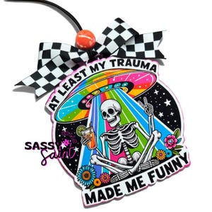 May include: A decorative ornament featuring a skeleton sitting in a lotus position, holding a drink under a colorful UFO. The text reads AT LEAST MY TRAUMA MADE ME FUNNY and SASSY SAINT. It is topped with a black and white checkered bow and orange bead.