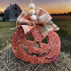 May include: A glittery orange jack-o'-lantern ornament featuring a cutout face, topped with a plaid fabric bow and three white wooden beads on a string. The text SASSY SAINT is printed in the lower right corner of the pumpkin-shaped decoration.