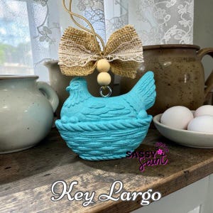 May include: A turquoise hen figurine in a woven basket, adorned with a burlap bow and wooden beads. The figurine is part of a farmhouse-style decor, with a rustic aesthetic. The text "Key Largo" is at the bottom.