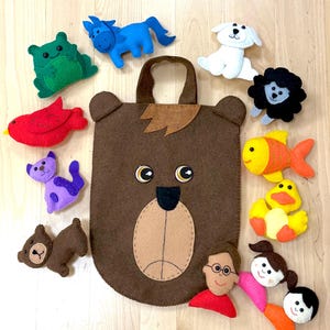 Può includere: Una borsa a mano a forma di orso in feltro marrone con una faccia da orso, circondata da figure di animali e personaggi in feltro. Le figure includono una rana verde, un cavallo blu, un cane bianco, un leone nero, un uccello rosso, un gatto viola, un orso marrone, un pesce giallo, un'anatra gialla e due persone.