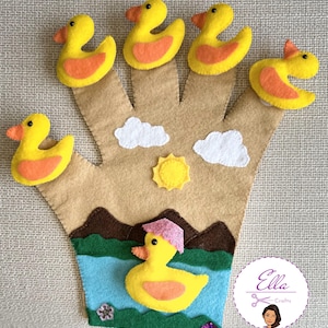 以下が含まれることがあります： 茶色の手のひらと黄色いアヒルの指人形が付いたフェルトの手人形。人形には、緑の草、青い水、茶色の山のある池に黄色いアヒルがいるシーンもあります。右下隅に「Ella Crafts」の文字が見えます。
