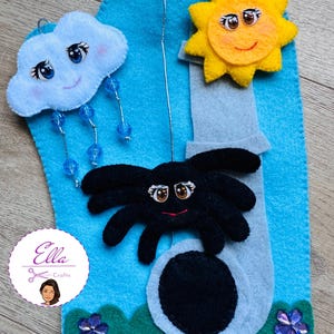 May include: A blue felt board with a felt sun, cloud, spider, and a hole for a hand puppet. The sun is yellow with a smiling face and the cloud is light blue with a smiling face. The spider is black with a smiling face and eight legs. The hole is gray and has a black circle in the center.