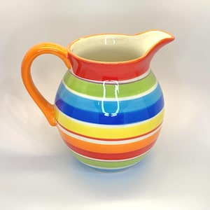 May include: A colorful striped ceramic pitcher with a bright orange handle. The pitcher has a red, orange, yellow, green, and blue striped design.