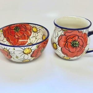 May include: A ceramic bowl and mug set. The bowl and mug are white with a floral pattern of red poppies and yellow-centered daisies. Both pieces have a blue rim. The mug has a blue handle.