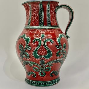 May include: A red ceramic pitcher with green and white floral designs. The pitcher has a wide mouth and a handle.