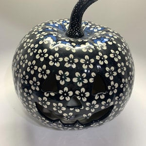 May include: A ceramic jack-o'-lantern with a dark blue background and white floral pattern. The pumpkin has a carved face with triangular eyes and a smiling mouth. The stem is dark blue with white speckles. A festive Halloween decoration.