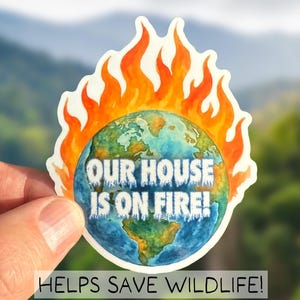 Könnte beinhalten: Ein Aufkleber mit einer Aquarellillustration der brennenden Erde. Der Text "OUR HOUSE IS ON FIRE!" steht über dem Globus, darunter "HELPS SAVE WILDLIFE!". Der Aufkleber wird vor einem verschwommenen natürlichen Hintergrund gehalten.