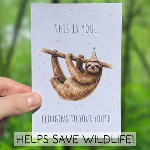 Peut inclure: Carte de vœux avec une illustration d'un paresseux portant un chapeau de fête, accroché à une branche. Le texte dit : « THIS IS YOU... CLINGING TO YOUR YOUTH » et « HELPS SAVE WILDLIFE ! » sur fond bleu clair.