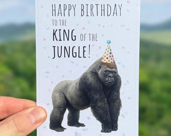 May include: A birthday card featuring a gorilla wearing a party hat. The card reads "Happy Birthday to the King of the Jungle!" and "Helps Save Gorillas!" in a white font. The background is a blurred outdoor scene.