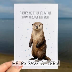May include: A greeting card featuring a watercolor illustration of an otter. The card reads "THERE'S NO OTTER I'D RATHER FLOAT THROUGH LIFE WITH" with small blue heart accents. The bottom of the card says "HELPS SAVE OTTERS!"