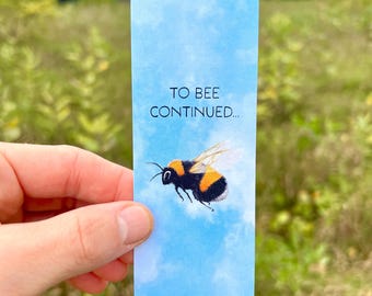 May include: A blue bookmark with a white cloud background and a bumble bee flying in the sky. The bookmark has the text "To Bee Continued..." printed on it.