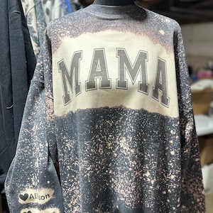 Puede incluir: Sudadera negra con un diseño salpicado de lejía blanca. La sudadera tiene la palabra "MAMA" impresa en grandes letras negras en el pecho. La manga izquierda tiene cuatro corazones negros con los nombres "Allison", "Waylon", "Aiden" y "Carson" impresos debajo de cada corazón.
