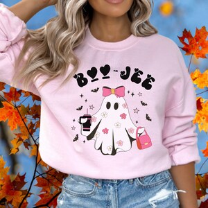 May include: A light pink crewneck sweatshirt featuring a graphic of a ghost wearing a pink bow, holding a dark drink and a pink handbag. The ghost is decorated with small white and pink flowers. The text BOO-JEE is printed in black above.