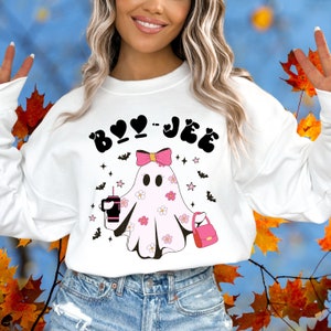 May include: A white long-sleeve sweatshirt featuring a graphic of a light pink ghost with a pink bow, floral patterns, a black tumbler, and a pink handbag. The text BOO-JEE is printed in black above the ghost, surrounded by small black bats and pink stars.