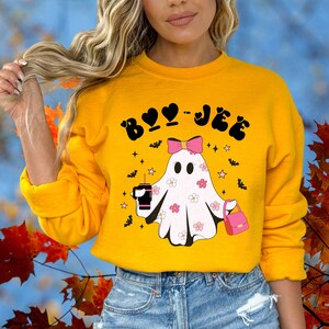 May include: A mustard yellow sweatshirt featuring a graphic of a white ghost with pink flowers, a pink bow, a pink handbag, and a black tumbler. The text "BOO-JEE" is printed in black above the ghost, surrounded by small black bats and pink stars.