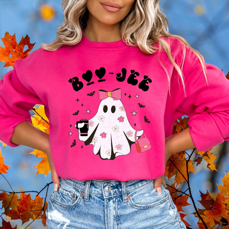 May include: A bright pink crewneck sweatshirt featuring a graphic of a white ghost with a pink bow, holding a black tumbler and a pink handbag. The ghost is decorated with small pink and white flowers. The text BOO-JEE is printed in black above.