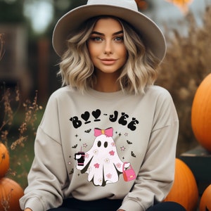 May include: A beige crewneck sweatshirt featuring a graphic of a white ghost with pink flowers, a pink bow, a pink handbag, and a black drink cup. The text "BOO-JEE" is printed in black above the ghost, surrounded by small black bats and pink stars.