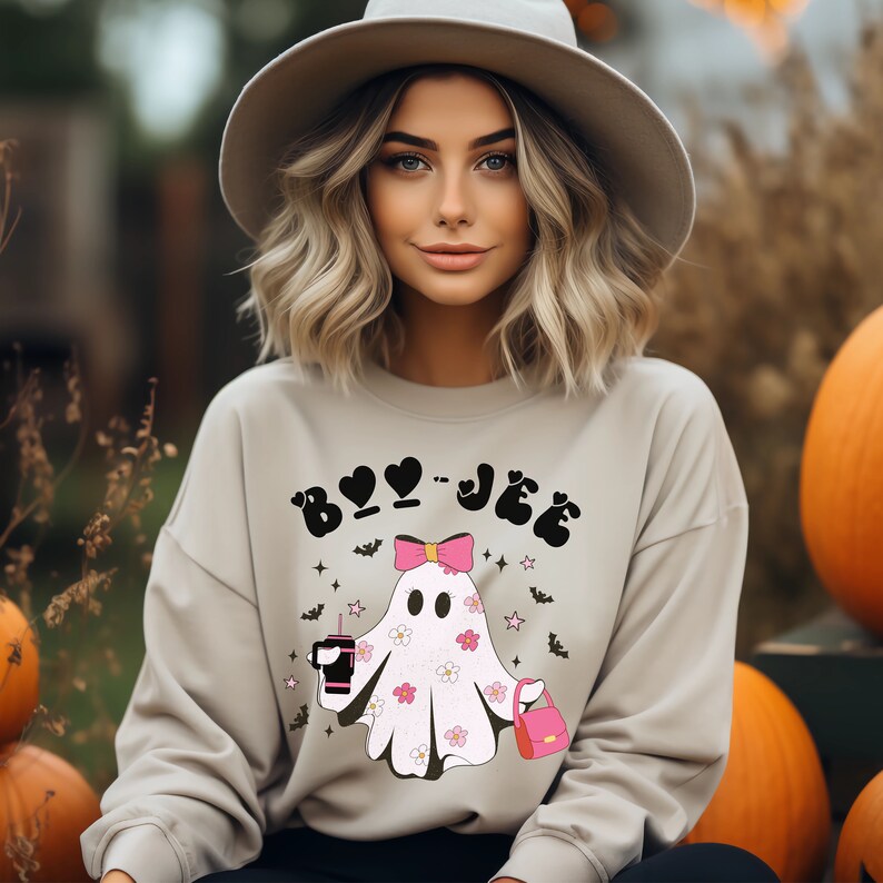 May include: A beige crewneck sweatshirt featuring a graphic of a white ghost with pink flowers, a pink bow, a pink handbag, and a black drink cup. The text "BOO-JEE" is printed in black above the ghost, surrounded by small black bats and pink stars.