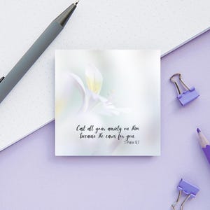 May include: A square white sticky note with a soft floral design and the text "Cast all your anxiety on Him because He cares for you. 1 Peter 5:7". A gray pen, purple pencil, and two purple binder clips are on a light purple surface.