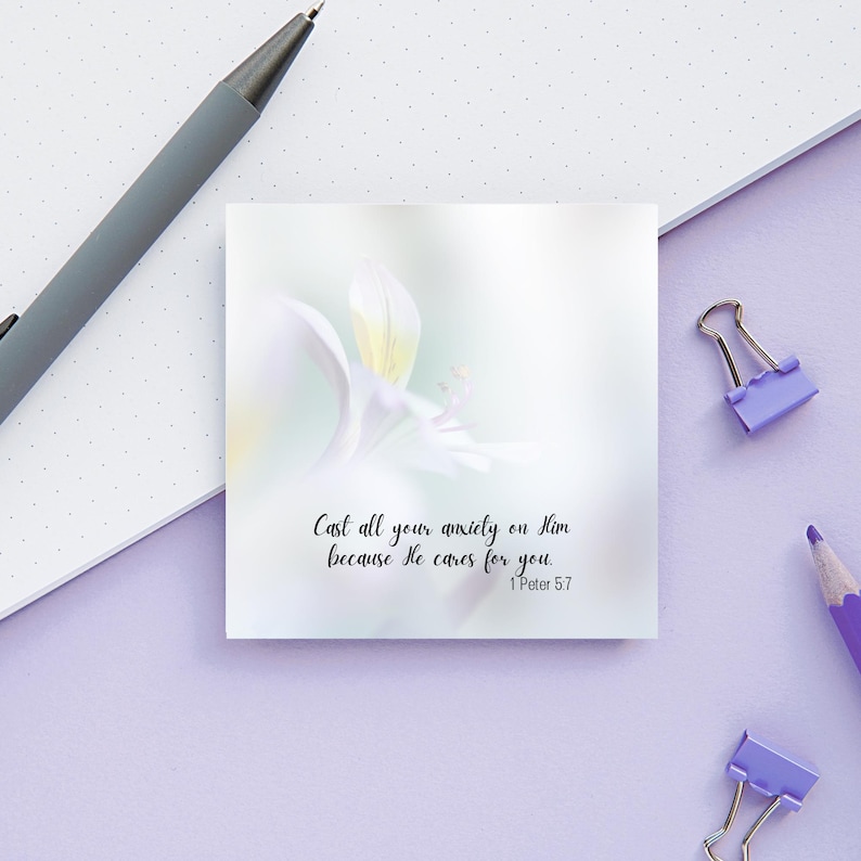May include: A square white sticky note with a soft floral design and the text "Cast all your anxiety on Him because He cares for you. 1 Peter 5:7". A gray pen, purple pencil, and two purple binder clips are on a light purple surface.