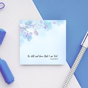 May include: A square, light blue sticky note with a floral design and the text "Be still and know that I am God. Psalm 46:10." A white pen and blue markers are in the background.