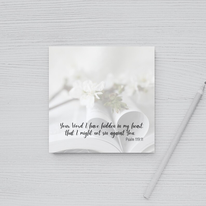 May include: A square, white card with the text "Your Word I have hidden in my heart, that I might not sin against You. Psalm 119:11" in black script. The card is placed on an open book with white flowers and a pencil.