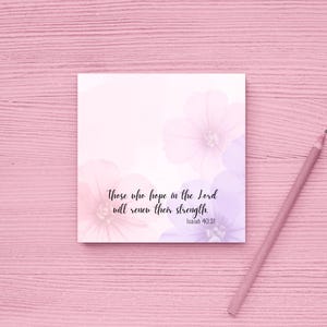 May include: A square, floral-themed notepad with the text "Those who hope in the Lord will renew their strength. Isaiah 40:31." The notepad features soft pink and purple watercolor flowers. A pink pencil rests on a pink wooden surface.