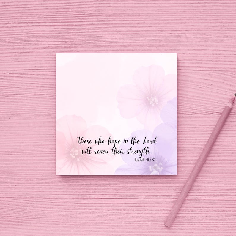 May include: A square, floral-themed notepad with the text "Those who hope in the Lord will renew their strength. Isaiah 40:31." The notepad features soft pink and purple watercolor flowers. A pink pencil rests on a pink wooden surface.
