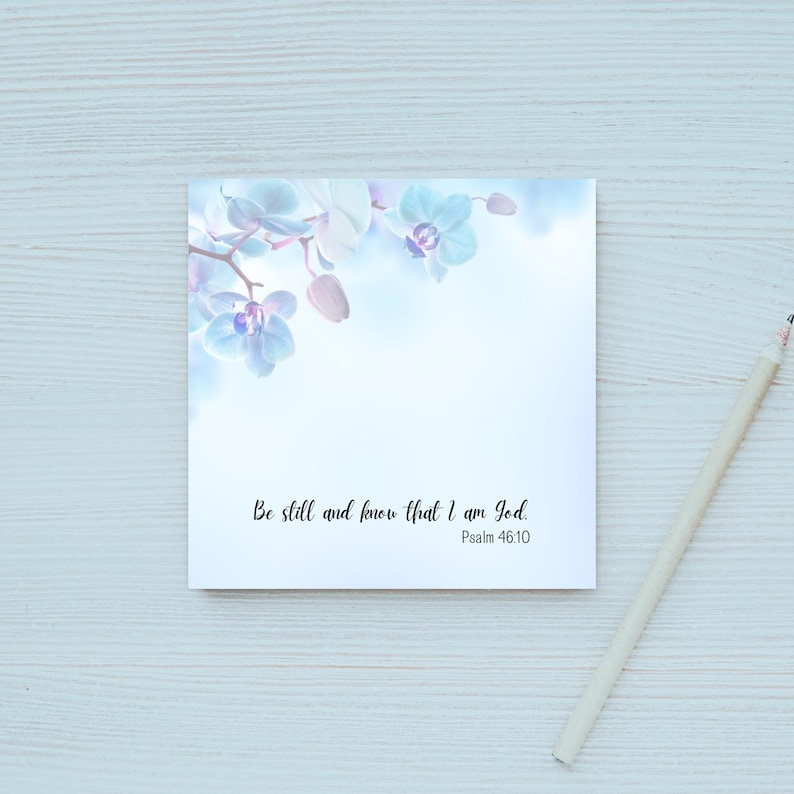 May include: A square notepad with a floral design featuring light blue orchids and the text "Be still and know that I am God. Psalm 46:10." A light-colored pencil rests on the side.