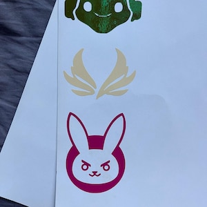 May include: Three Overwatch character decals on a white background. The top decal is green and features a robot with headphones. The middle decal is gold and features a pair of wings. The bottom decal is pink and features a bunny rabbit with angry eyes.
