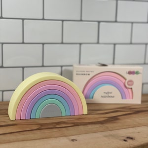 Puede incluir: Un juguete apilable de arcoíris en colores pastel con una capa superior de color amarillo claro, capas rosas, moradas, verdes y grises. El juguete está sobre una superficie de madera, con una caja en el fondo que dice "my first rainbow."