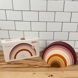 Puede incluir: Un juguete de arcoíris apilable de silicona en tonos marrón, rosa y naranja. El juguete se muestra junto a su embalaje, que dice "my first rainbow". El juguete está diseñado para niños de 3 meses en adelante.