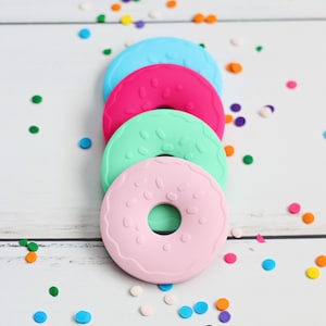 Puede incluir: Cuatro moldes de silicona con forma de rosquilla en tonos pastel rosa, verde, azul y fucsia. Cada molde tiene un agujero en el centro y pequeñas chispas blancas. Los moldes están dispuestos en una pila vertical sobre una superficie de madera blanca, con confeti colorido esparcido.