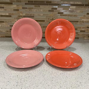 May include: A set of four ceramic plates featuring a glossy finish and concentric ring detailing. Two plates are a soft pastel pink, while the other two are a vibrant, saturated orange. Each plate has a raised rim and a smooth, circular center.