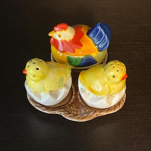 May include: A ceramic hen-shaped container with a red comb, yellow, blue, and orange body, and two yellow chick salt and pepper shakers in eggshell halves. The set sits on a brown, leaf-shaped tray.
