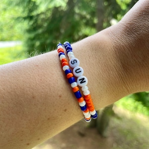 May include: Two beaded bracelets, one orange, white, and blue, and the other white and blue, with the word "SUN" spelled out in white beads.