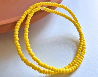 May include: A circular necklace composed of small, uniform, bright yellow seed beads strung together. The beads have a smooth, glossy finish and are arranged in a single continuous strand that forms a simple, rounded loop shape.