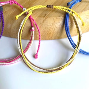 May include: Three colorful friendship bracelets made of woven string. The bracelets are in shades of pink, yellow, and blue.