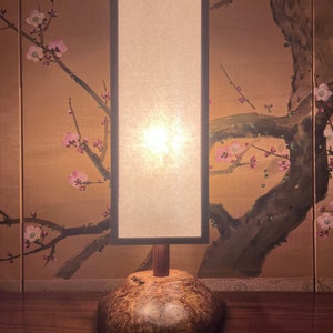 May include: A modern table lamp with a wooden base and a rectangular white lampshade. The lamp is lit and casting a warm glow. The lamp is sitting on a wooden table.