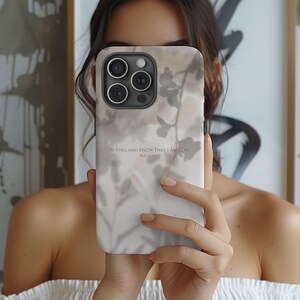 May include: A phone case featuring a soft, neutral-toned background with a subtle, blurred botanical shadow pattern. The text "BE STILL AND KNOW THAT I AM GOD. PSALM 46:10" is printed in a clean, dark font across the centre of the case.