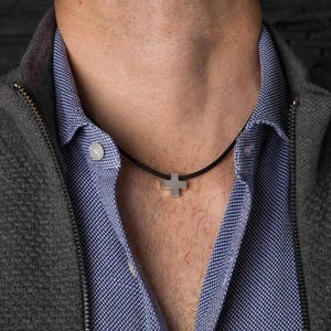 Peut inclure: Un collier avec un pendentif en forme de croix argentée sur un cordon noir. La croix est de conception simple et minimaliste. Le collier est porté sur une chemise bleue à motifs et une veste grise zippée.