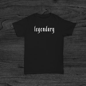 Puede incluir: Camiseta negra con la palabra "legendario" impresa en blanco en una fuente gótica.